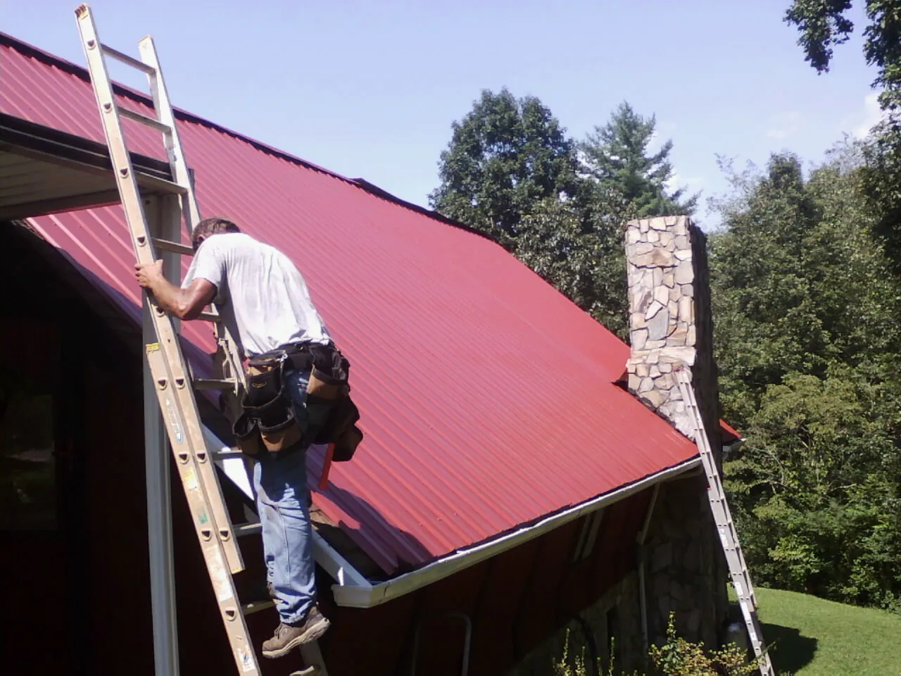 Roofer on a ladder beside a red metal roof and stone chimney — Soffit & Fascia Repair by Roofing Brothers in Brush